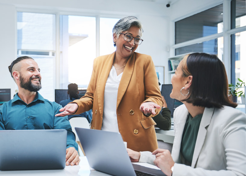Smiling woman standing and shrugging while two coworkers smile up at her while working at their laptops.