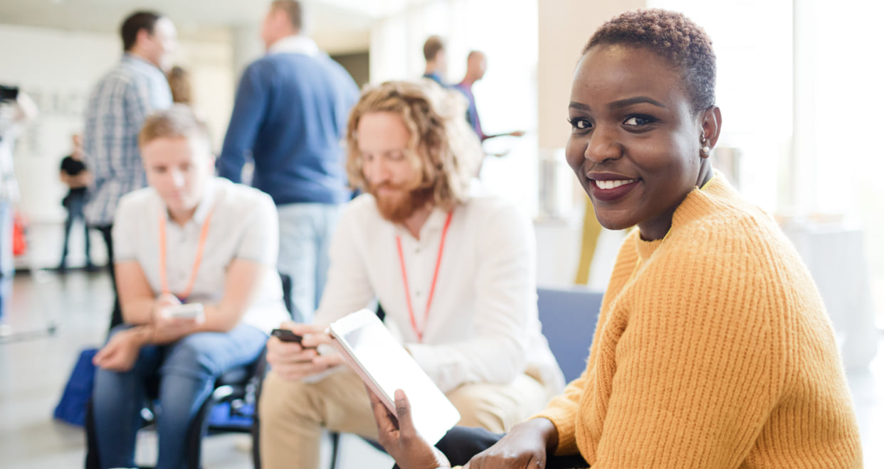 Woman Smiling while holding a tablet with coworkers are chatting in the background.