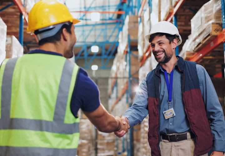 Two coworkers wearing hard hats and shaking hands in a warehouse setting.