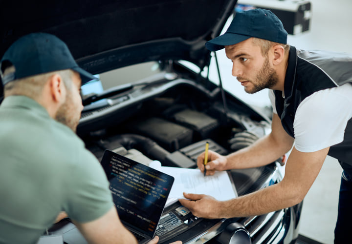 Two mechanics looking inside of a hood of a car while writing notes on a report.