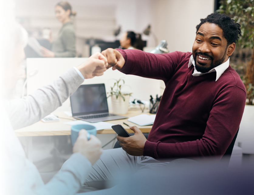 Two smiling coworkers fist bumping while sitting at their desks.