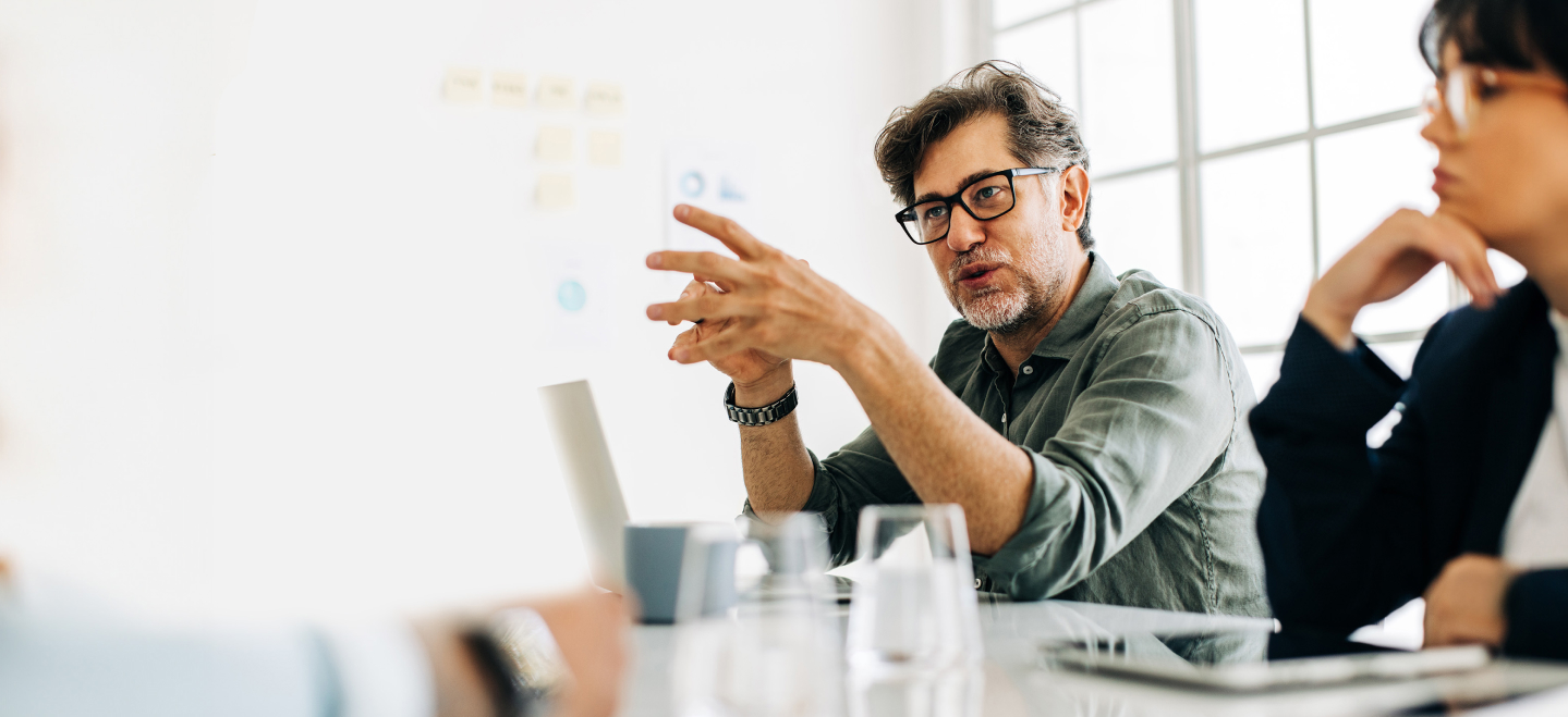 Man speaking animatedly while working at a table with his coworkers. A 53 out of 60 scale and an orange ‘Challenge the Process’ leadership behavior banner are in the bottom right corner.