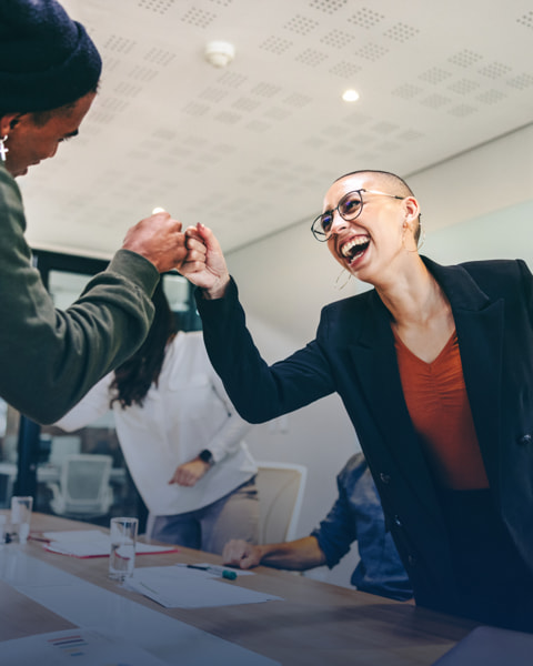 Two coworkers smiling and fist bumping across a meeting room table