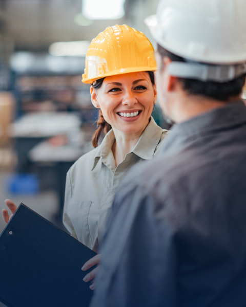 Two coworkers smiling while wearing hardhats in a warehouse setting