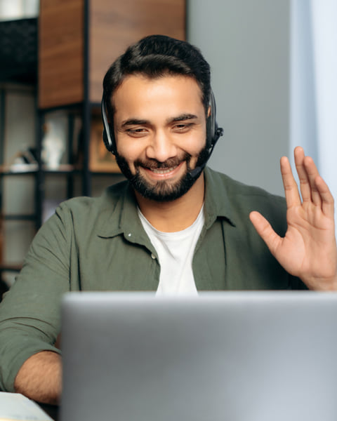 Smiling man wearing a headset and waving into a computer.