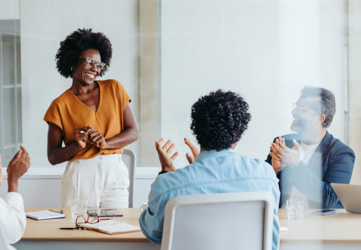 Woman standing in a meeting room while her coworkers clap for her.