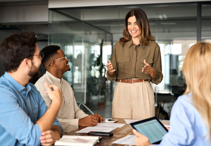 Woman standing and presenting information while her coworkers smile and listen.