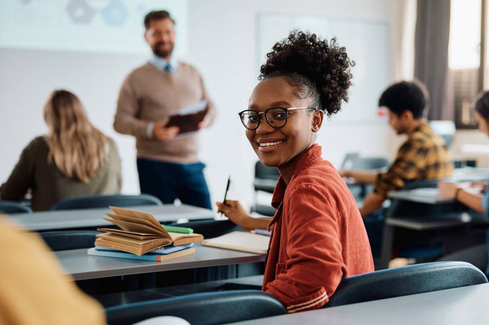 Smiling woman sitting at a desk in a classroom setting with a teacher presenting something in the background.