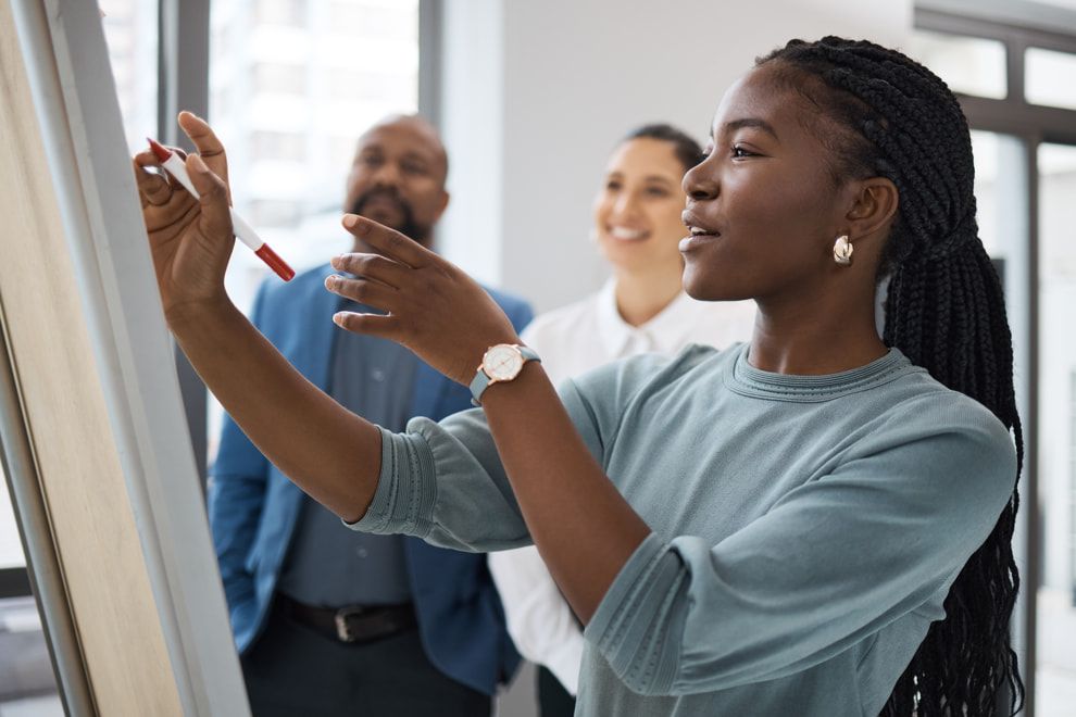 Woman writing something on a board, while two coworkers smile and look at her work