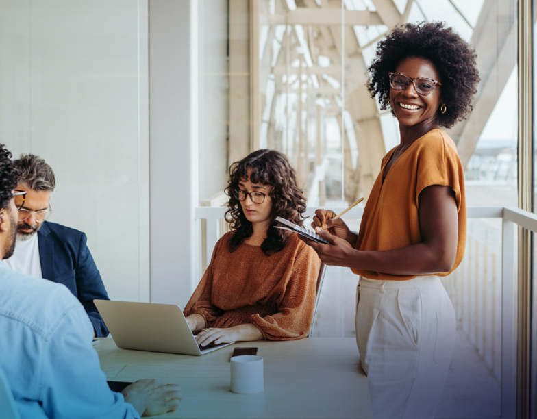 Standing woman writing something in a notebook, while coworkers sit at a table in the background.