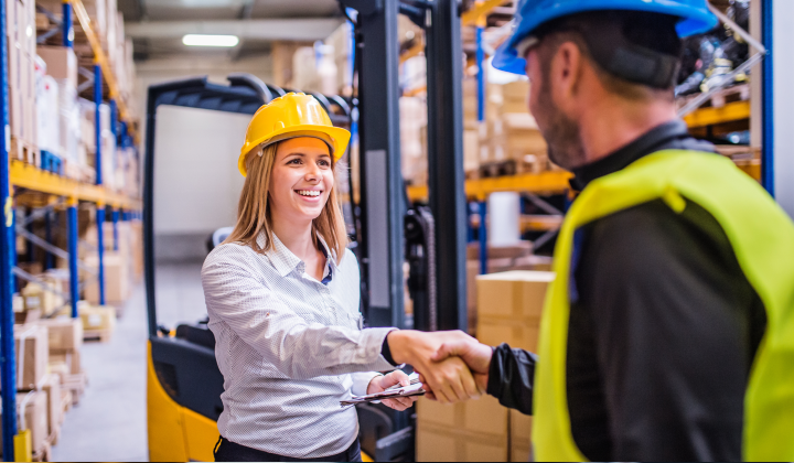Woman wearing a hard hat shaking hands with a man in a hard hat and yellow safety vest, in a warehouse setting.