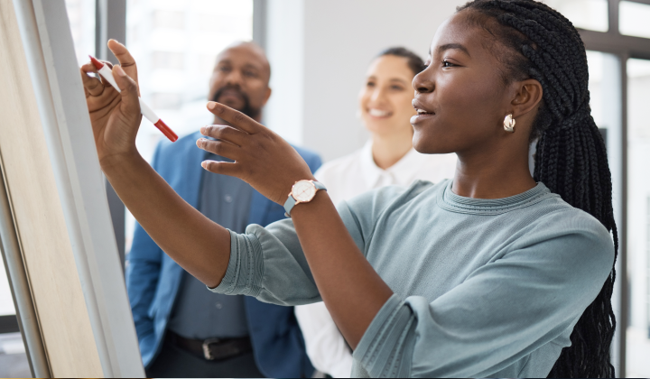 Woman writing on a large board while her coworkers smile in the background.