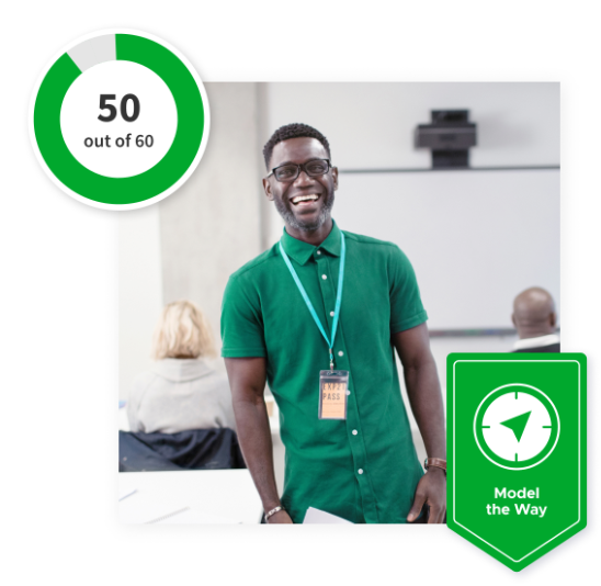 Teacher smiling in the middle of a classroom with a green ‘Model the Way’ banner at the bottom and a 50 out of 60 scale in the top left corner.